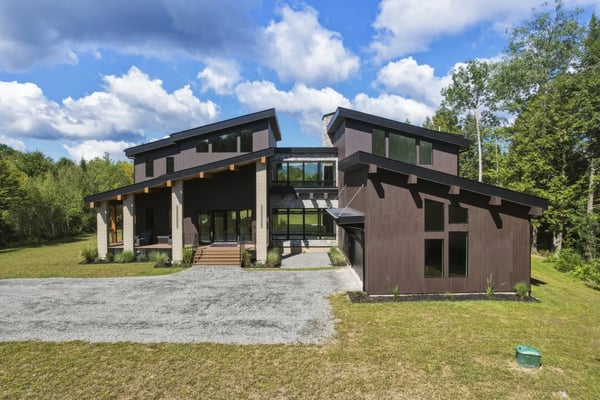 Modern two-story house with dark brown and white exterior, large windows, covered porch, and gravel driveway surrounded by green lawn and trees.