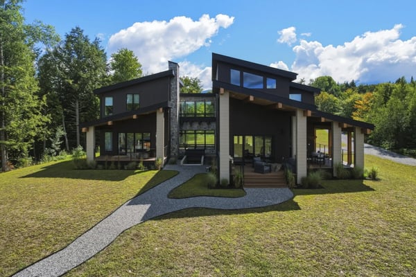 Modern black and white house with large windows, stone chimney, and covered porch surrounded by green lawn and forest.