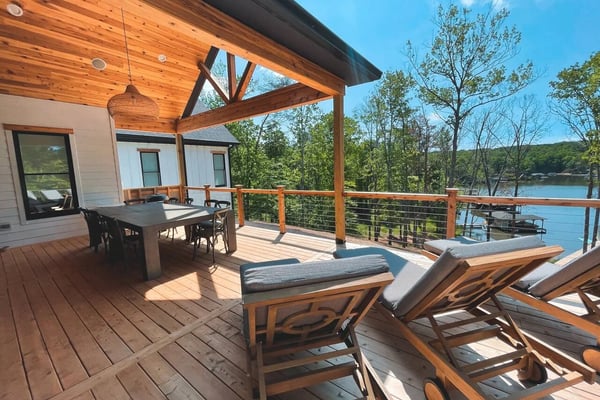 Wooden deck with lounge chairs and dining table overlooking a lake, with forested hills in background