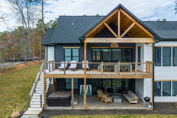 Modern two-story house with white and dark siding, wooden beam gable, multi-level decks with lounge chairs and hot tub, surrounded by bare trees and lawn.