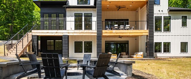 Modern two-story house with black and white exterior, wooden deck, and patio with Adirondack chairs surrounding fire pit.