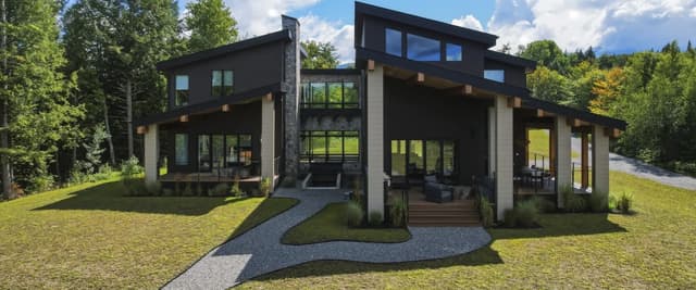 Modern black and white house with large windows, gravel pathway, and manicured lawn surrounded by forest and mountains.