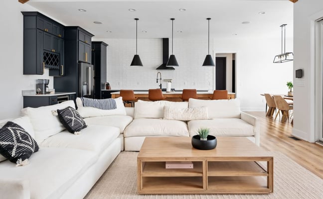 Open-plan living room with cream sectional sofa, wood coffee table, black kitchen cabinetry, and pendant lights overhead