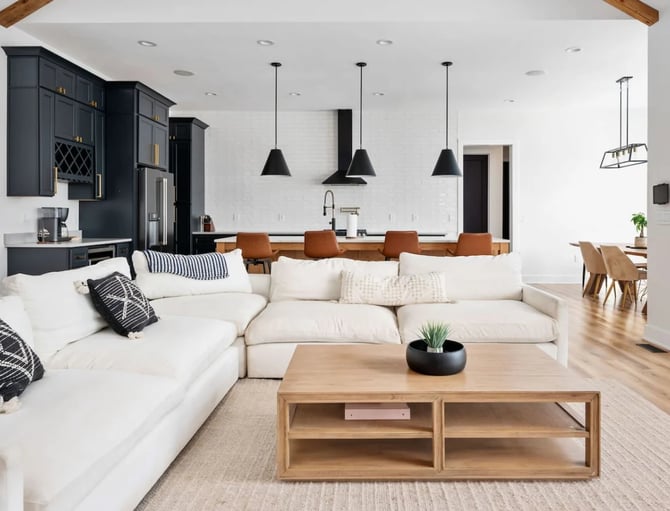 Open-plan living room with white sectional sofa, wood coffee table, black kitchen cabinets, and pendant lights overhead